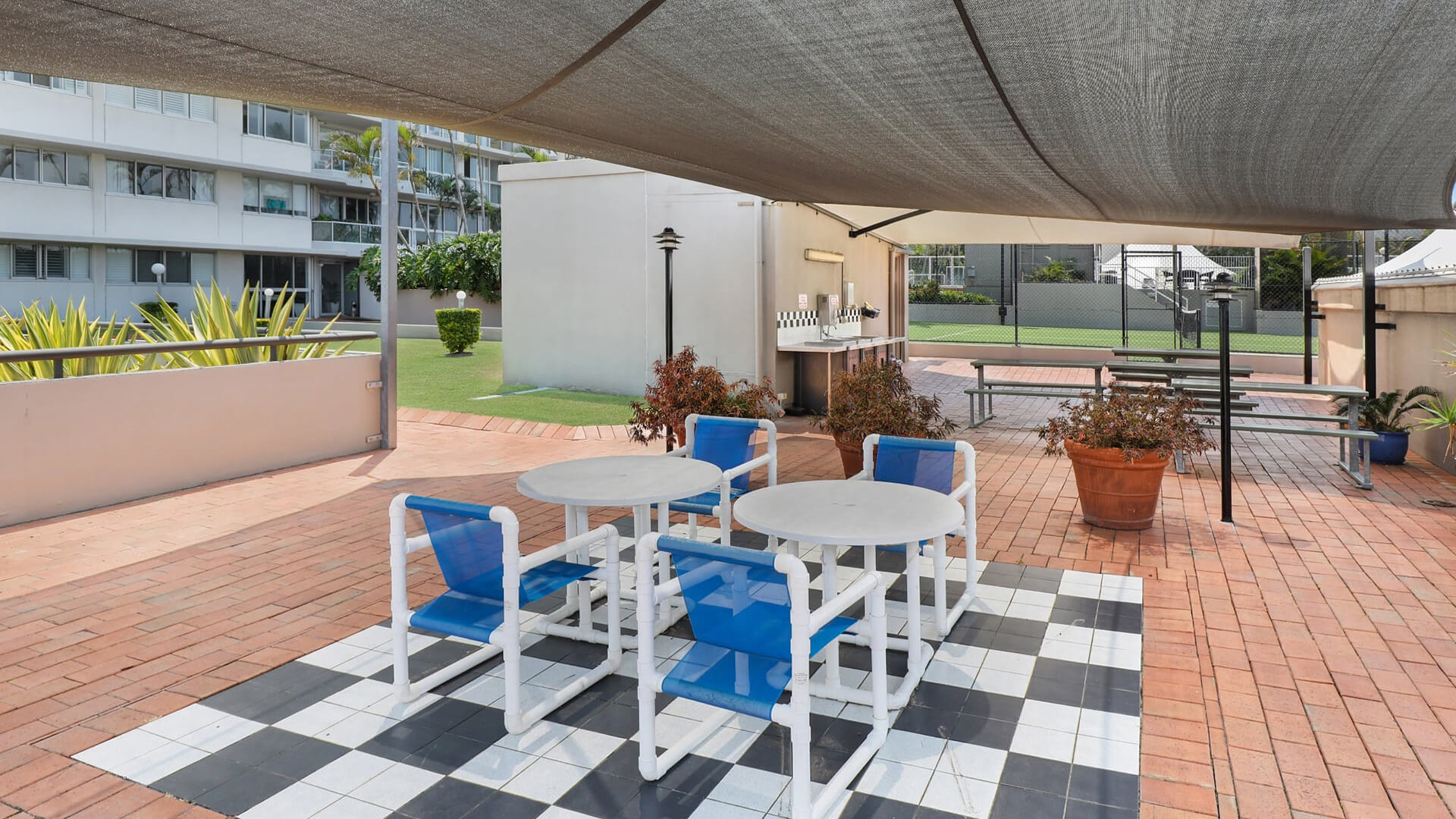 Shaded outdoor BBQ and seating area with blue-and-white chairs, tiled flooring, and nearby picnic tables overlooking the tennis court at ULTIQA Points North.