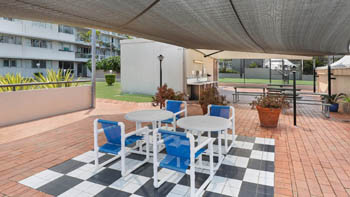 Shaded outdoor BBQ and seating area with blue-and-white chairs, tiled flooring, and nearby picnic tables overlooking the tennis court at ULTIQA Points North.