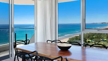 Close-up of the dining area, featuring a wooden table, black designer chairs, and sweeping views of the coastline and turquoise ocean through expansive glass windows.