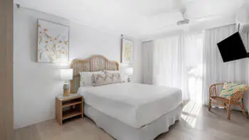 Coastal-style featuring a queen bed with rattan headboard, neutral tones, natural light, and a wall-mounted TV.