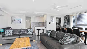 Living room area with a black leather couch, blue rug, wooden coffee table and kitchen in the background at ULTIQA Beach Haven on Broadbeach.