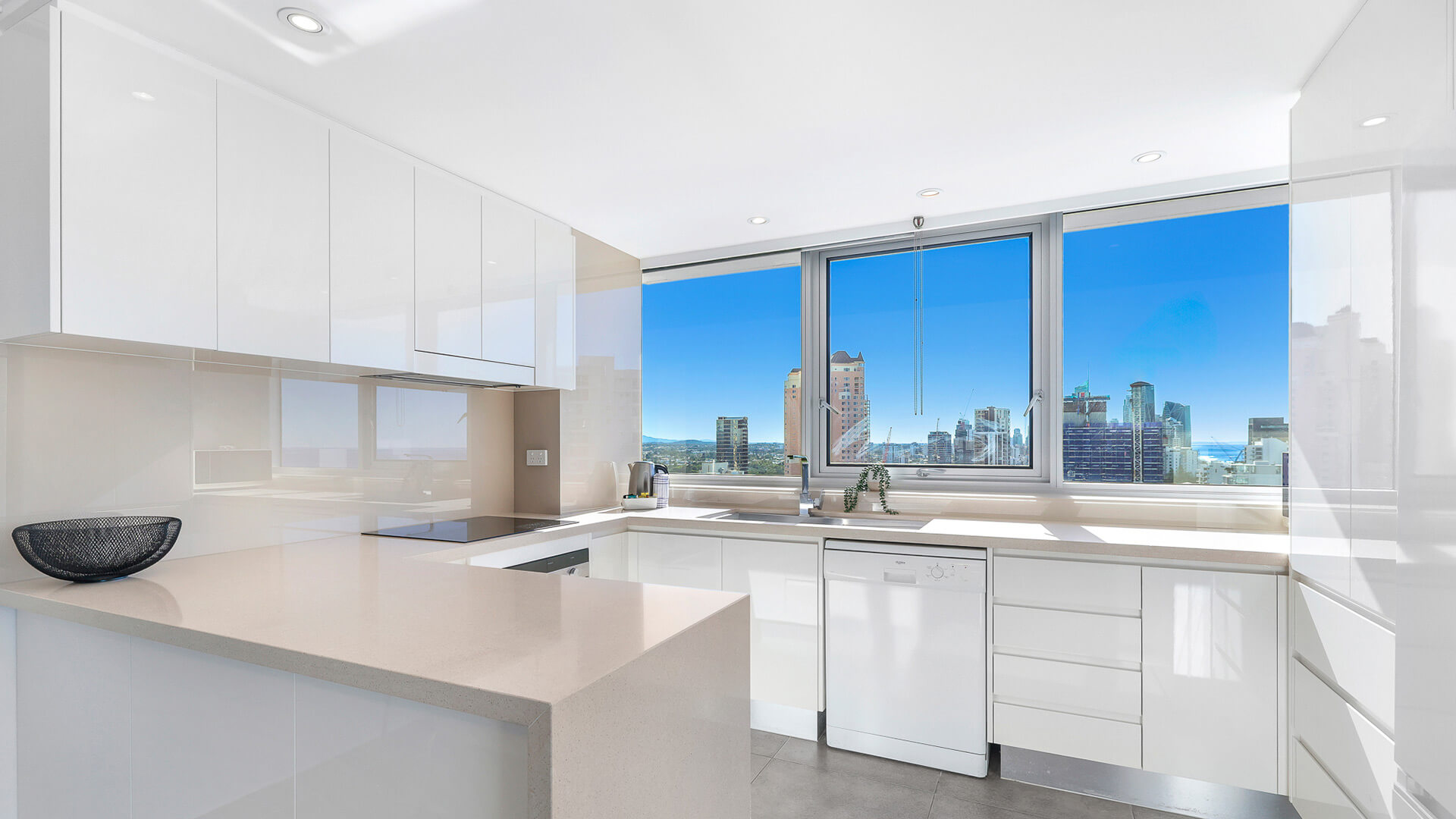 Modern fully equipped kitchen in the 2 Bedroom Ocean View Apartment at ULTIQA Beach Haven featuring sleek white cabinetry, stone benchtops, and city skyline views through large windows.