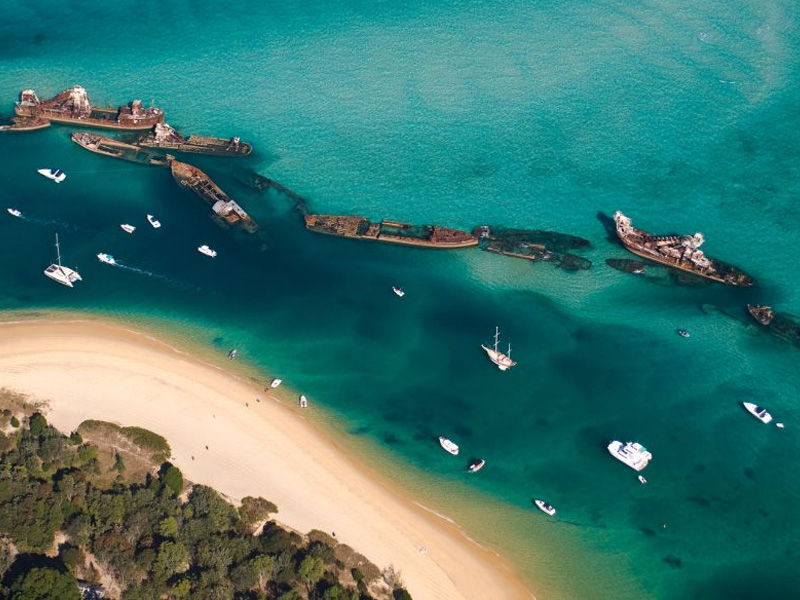 Aerial view of the Tangalooma Wrecks in turquoise waters, with snorkelers swimming around the partially submerged, rusted ship hulls near Moreton Island, Queensland.