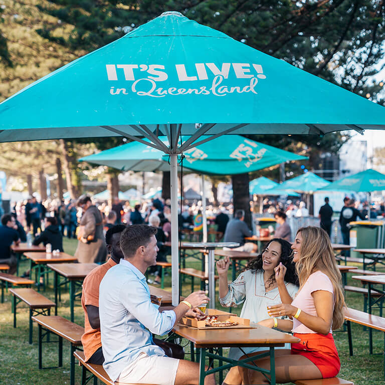 Friends enjoying food and drinks at an outdoor event in Queensland under a turquoise It's Live! in Queensland umbrella.