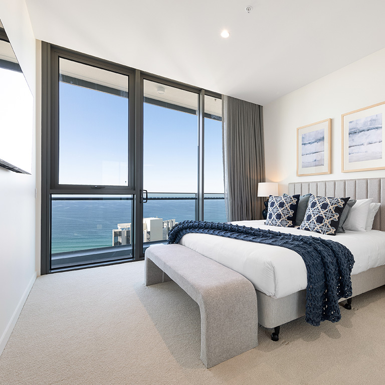 Ocean-view bedroom at ULTIQA Signature at Broadbeach, featuring a king bed with navy accents, floor-to-ceiling windows, wall-mounted TV, and modern coastal décor