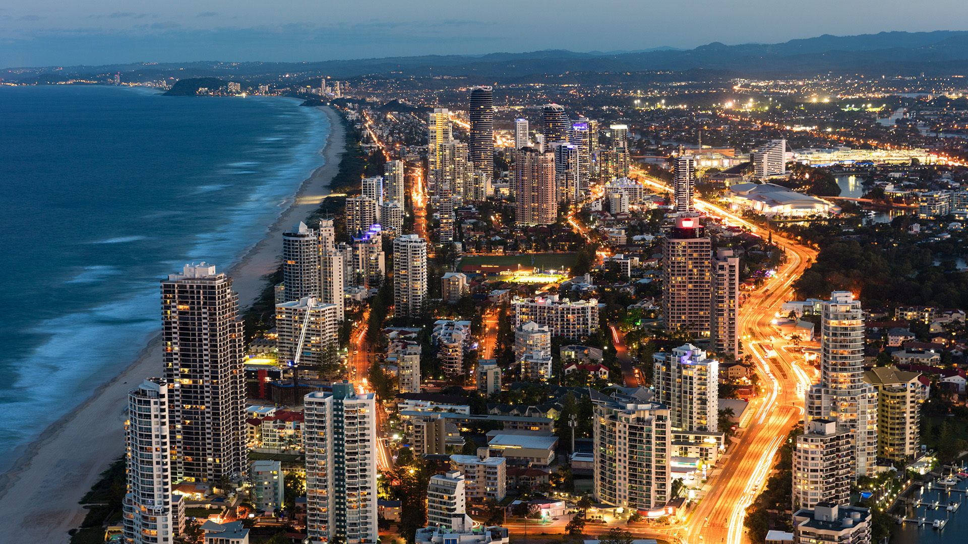 Aerial view of the Gold Coast skyline at dusk, showcasing illuminated high-rise buildings, city lights, coastal roads, and the stretch of beach meeting the ocean
