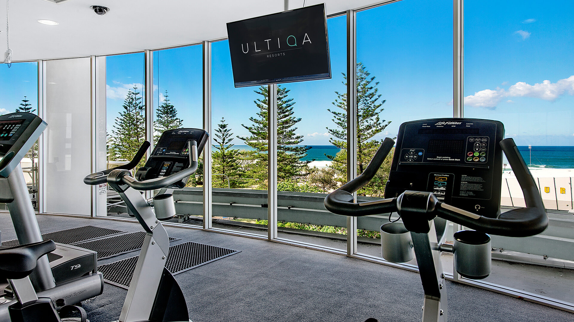 Ocean-view fitness room at ULTIQA Air on Broadbeach with stationary bikes, floor mats, and large windows overlooking pine trees and the ocean.