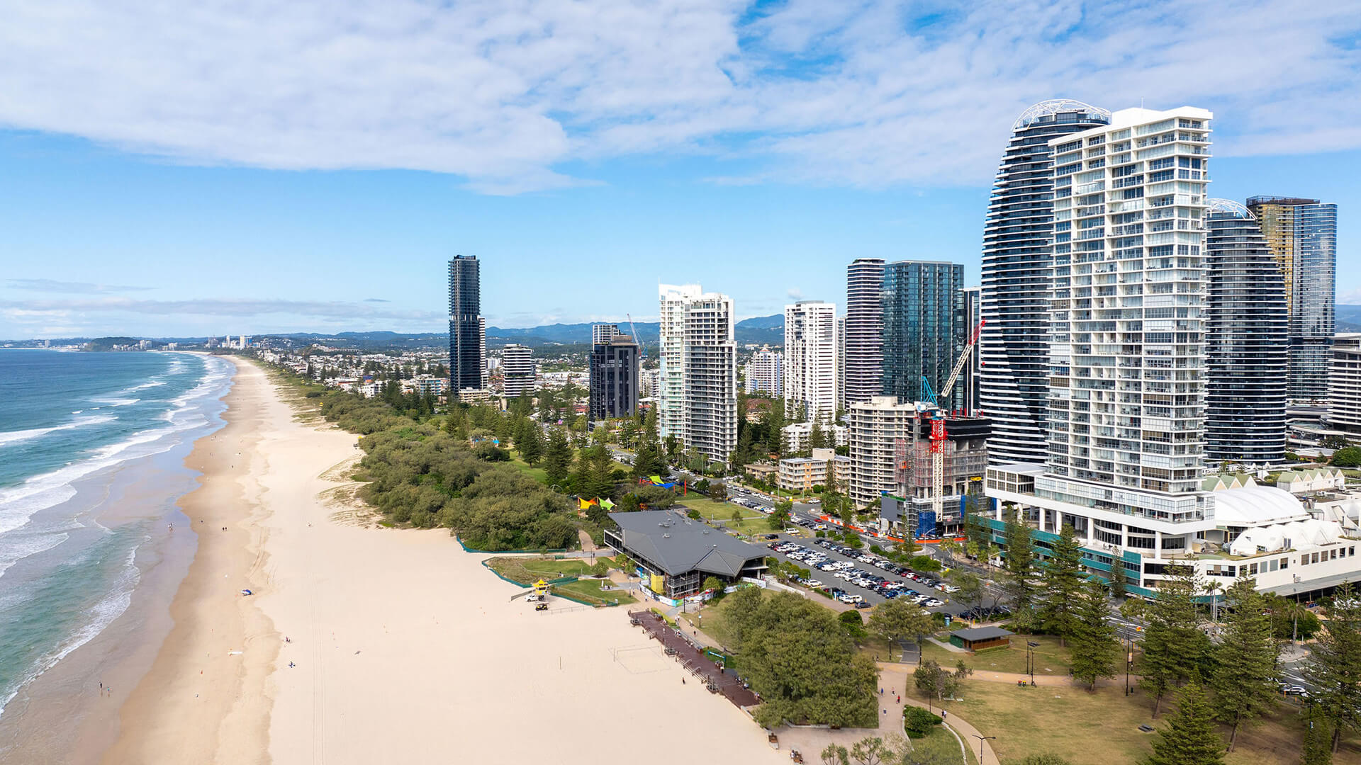 Coastal city view from ULTIQA Air on Broadbeach featuring high-rises, parklands, a beach with lifeguard station, and a construction site under a sunny sky.