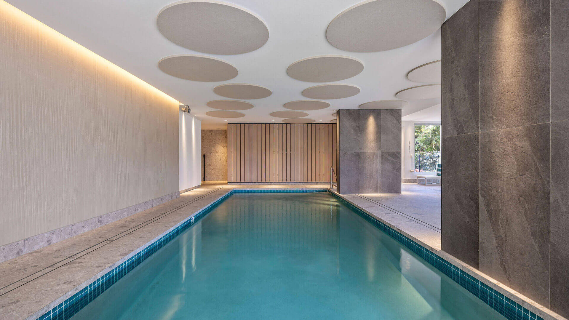 Indoor heated lap pool at ULTIQA Beach Haven, featuring elegant stone finishes, circular ceiling panels, and ambient lighting — perfect for a relaxing swim year-round.