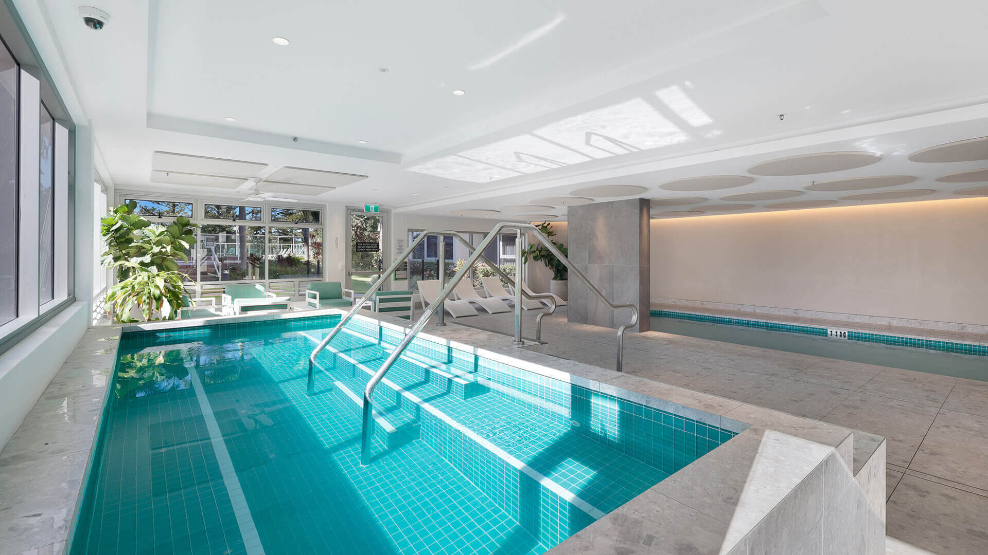 Indoor heated spa and lap pool area at ULTIQA Beach, featuring tiled steps, natural light, poolside seating and leafy surrounds - perfect for year-round relaxation.
