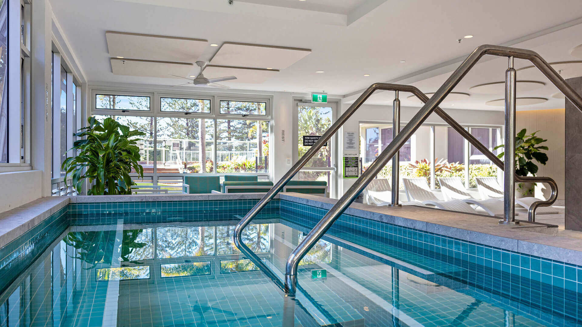 Indoor spa at ULTIQA Beach Haven with stainless steel handrails, natural light streaming through large windows, poolside lounge chairs and lush greenery for a relaxing retreat.
