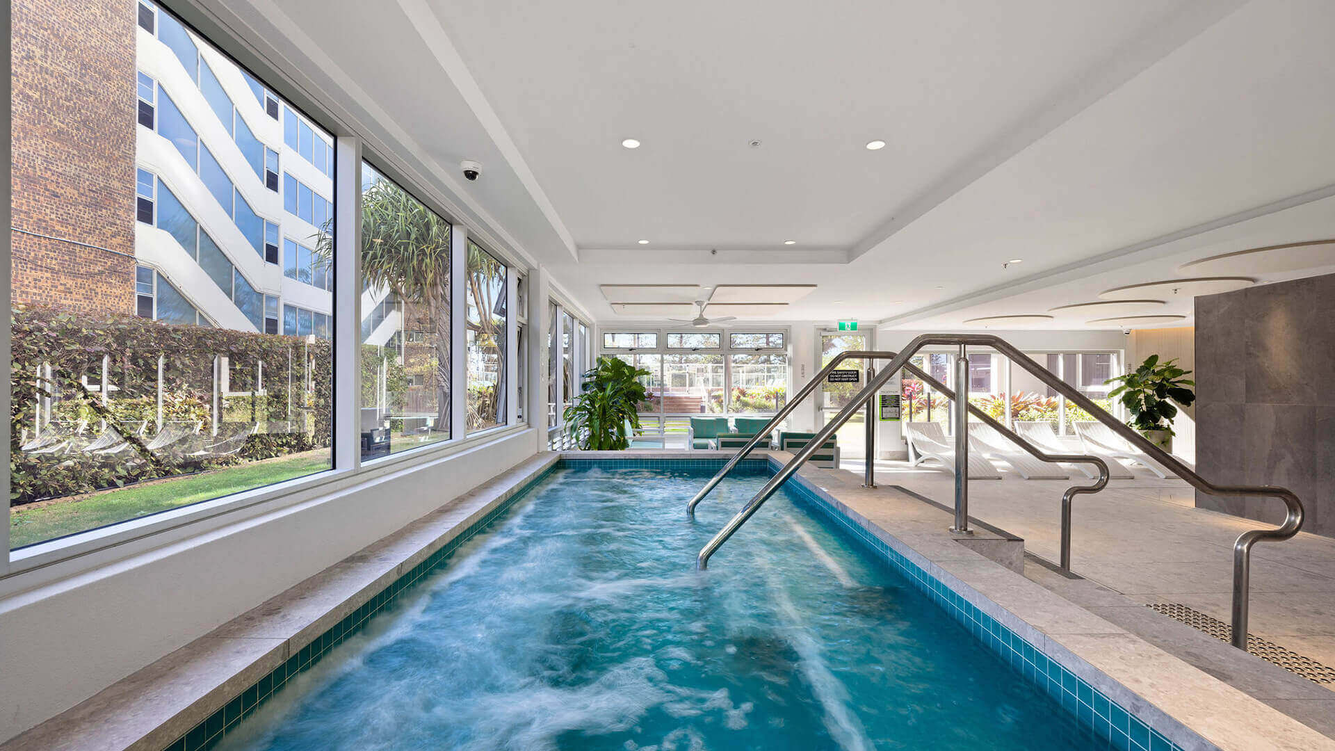 Indoor spa with bubbling jets at ULTIQA Beach Haven, featuring floor-to-ceiling windows, stainless steel handrails and poolside lounge seating for ultimate relaxation.