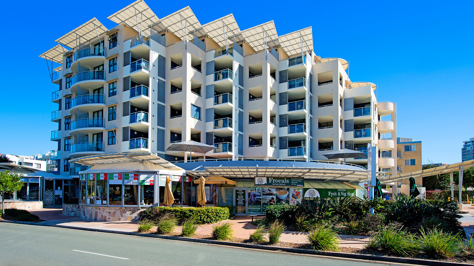 Street view of ULTIQA Shearwater Resort with balconies, ground-floor shops, and a bright blue sky backdrop.