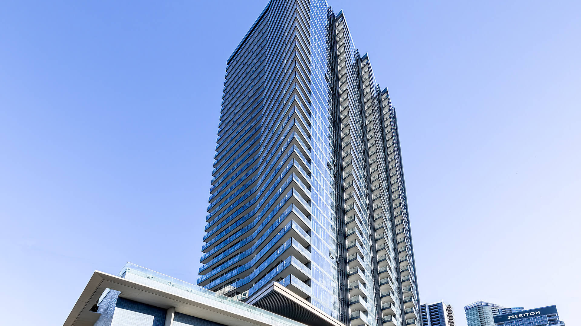 Tall, modern high-rise building with sleek glass facades and multiple balconies, set against a clear blue sky.