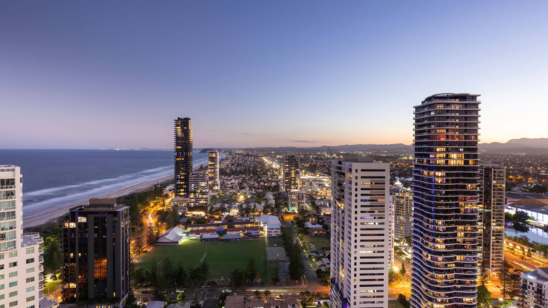 Cityscape view at twilight with high-rise buildings illuminated, a sports field in the foreground and the coastline stretching along the left side.