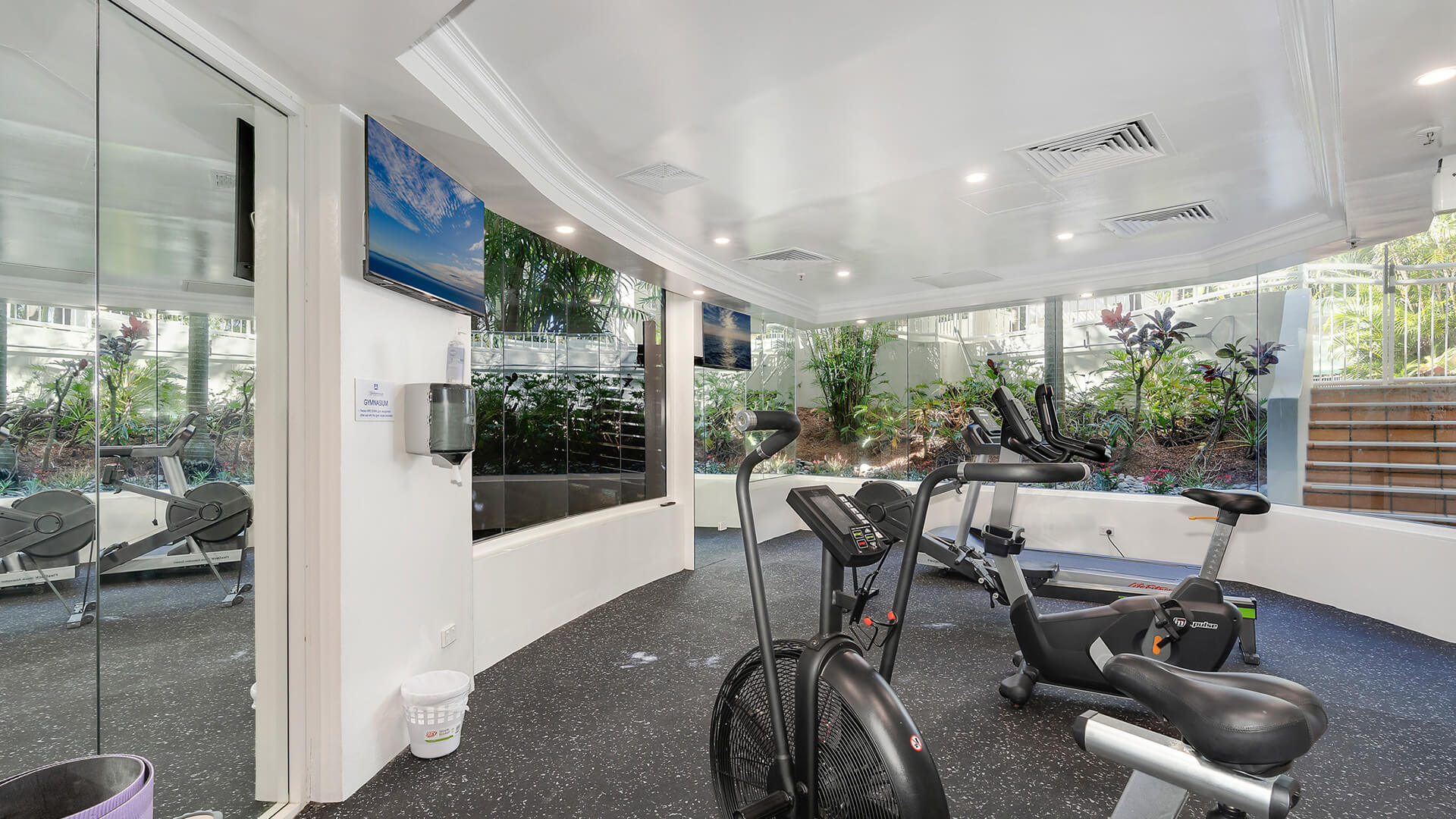 Fully equipped fitness centre at ULTIQA Burleigh Mediterranean Resort, featuring cardio machines, exercise bikes and views of lush garden landscaping through large windows.