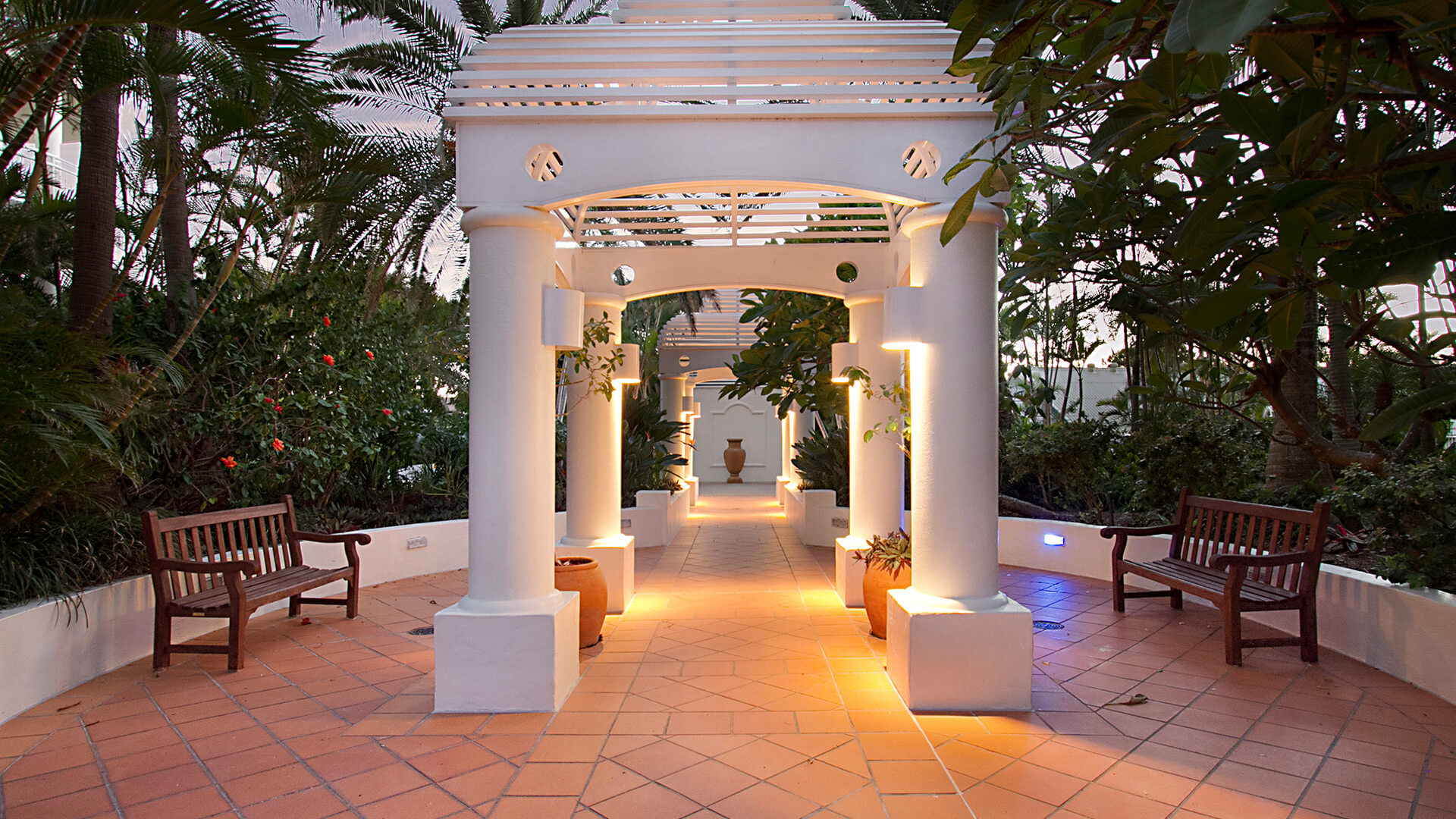 Illuminated Mediterranean-style walkway at ULTIQA Burleigh Mediterranean Resort, surrounded by lush tropical gardens and lined with wooden benches for a peaceful retreat.