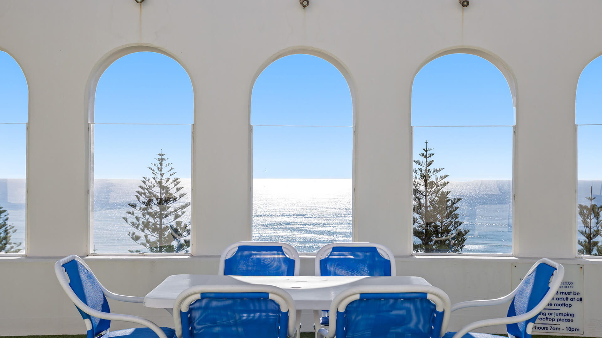 Rooftop dining area at ULTIQA Burleigh Mediterranean Resort, featuring ocean-view seating framed by arched windows and vibrant blue chairs.