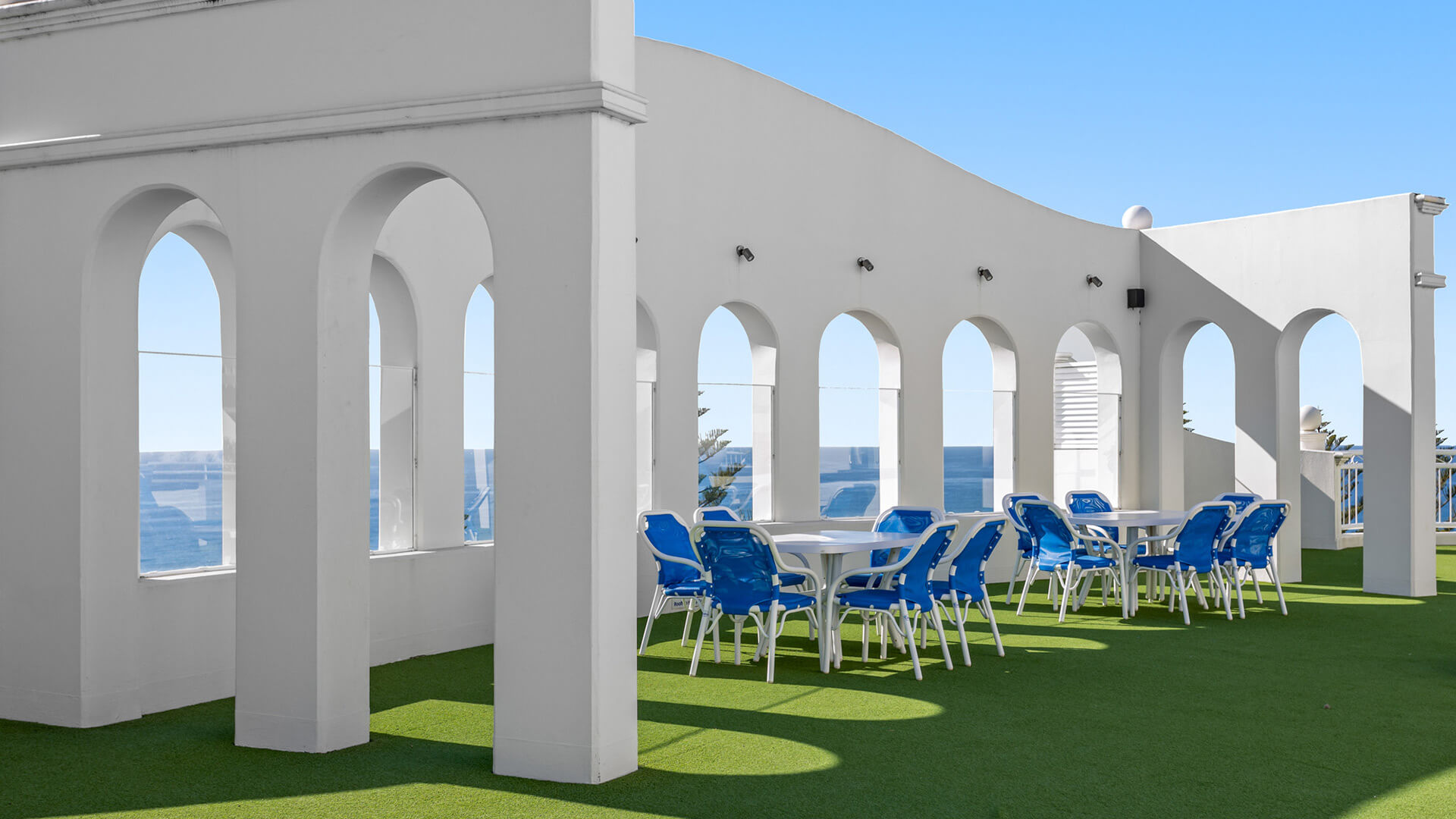 Rooftop terrace at ULTIQA Burleigh Mediterranean Resort, showcasing arched architecture, green turf flooring and multiple outdoor dining tables with blue seating overlooking the ocean.