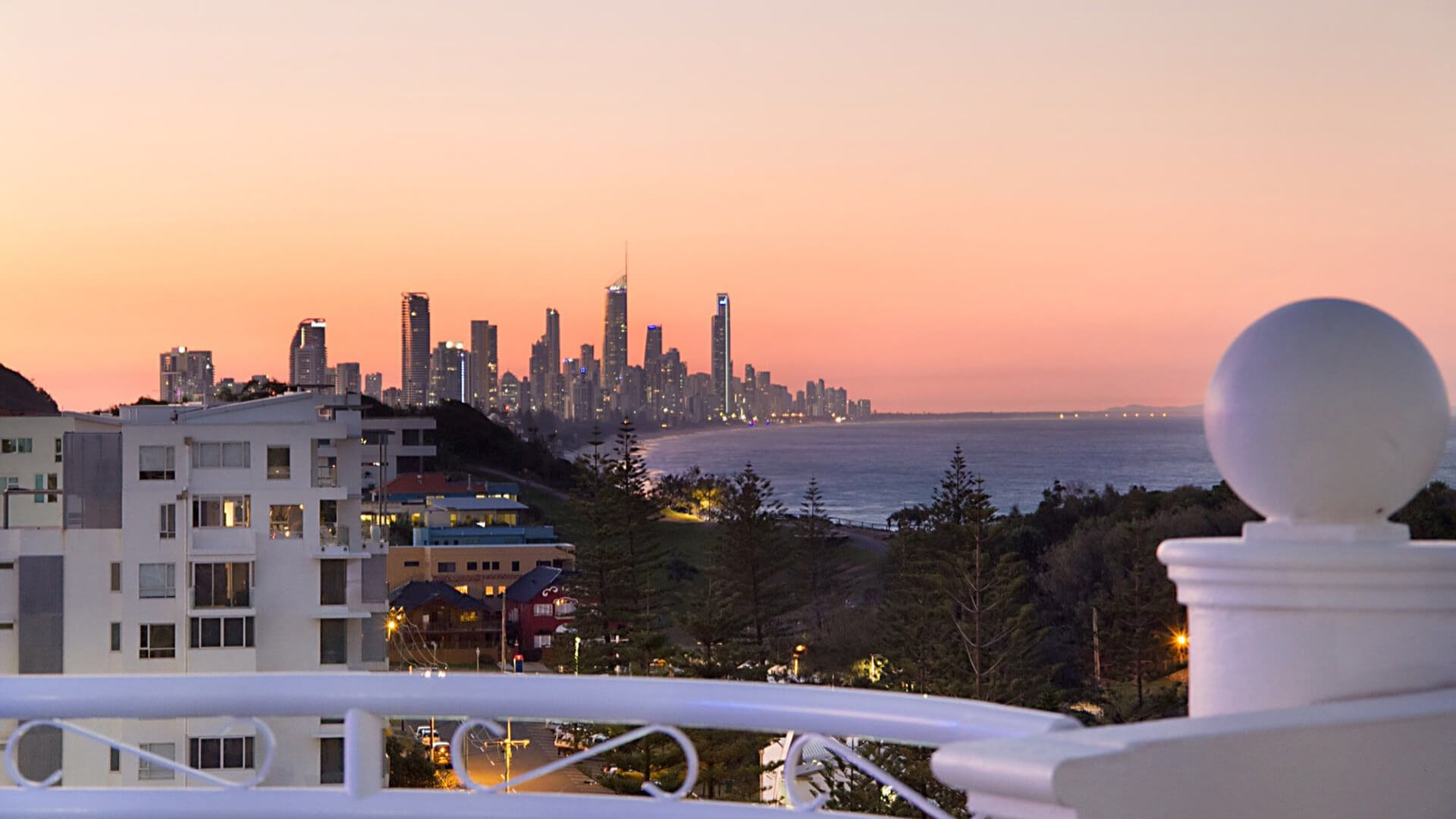 View from ULTIQA Burleigh Mediterranean Resort at sunset, showcasing the Gold Coast skyline, ocean, and surrounding buildings, with the warm glow of twilight creating a picturesque backdrop.