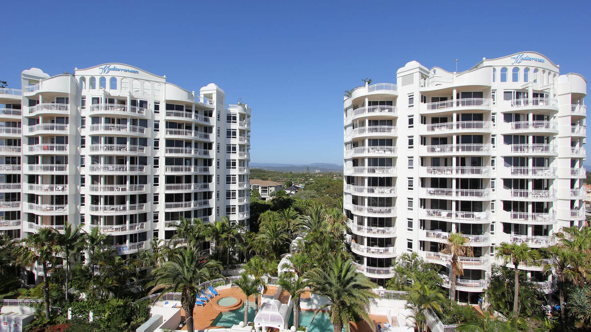 The twin towers of ULTIQA Burleigh Mediterranean Resort, framed by lush tropical gardens and featuring a central pool area with sun loungers, spa and shaded seating.