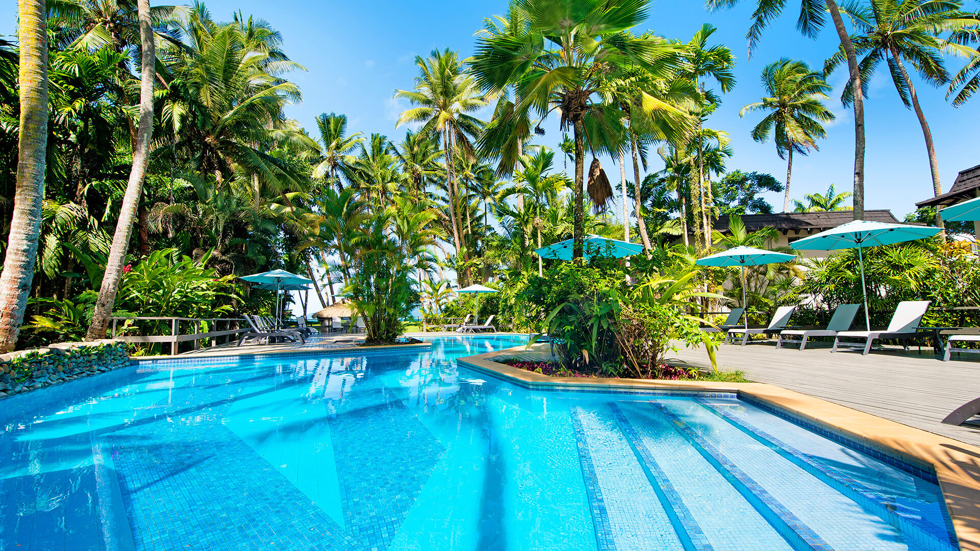 Resort-style swimming pool at ULTIQA Fiji surrounded by tropical gardens, palm trees and sun loungers with umbrellas.