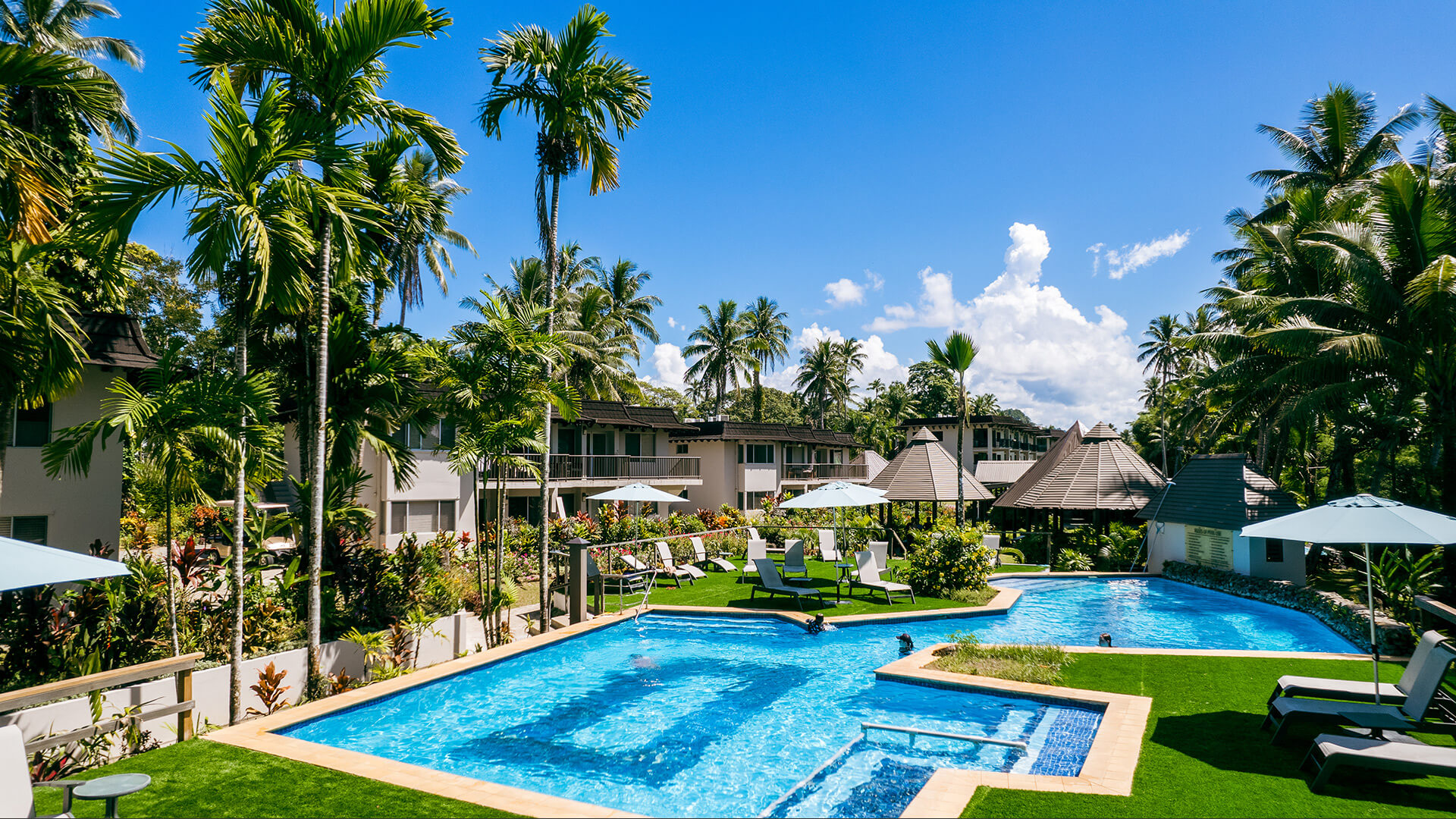 Resort pool area at ULTIQA Fiji with sun loungers, umbrellas, tropical gardens and surrounding accommodation buildings.