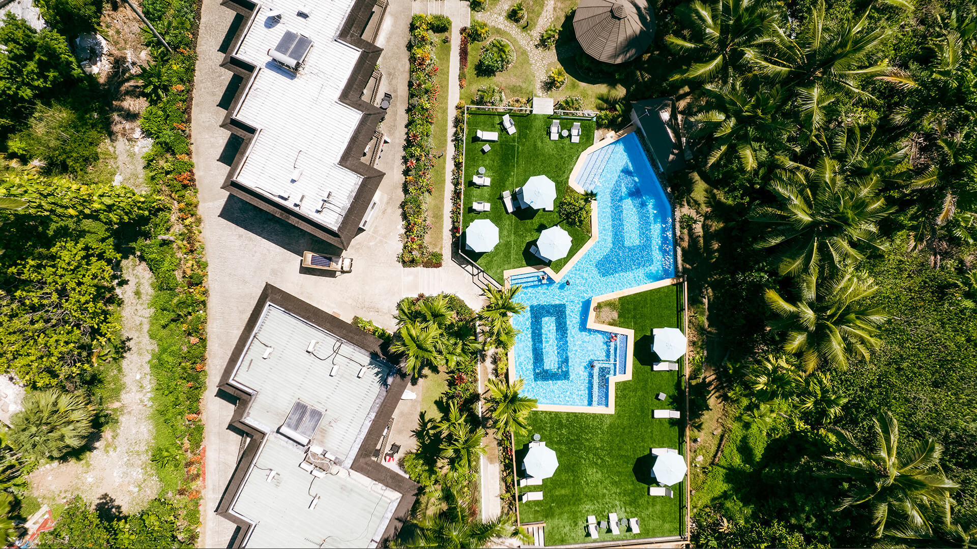 Aerial view of ULTIQA Fiji resort pool area with surrounding gardens, sun loungers and tropical palm trees.