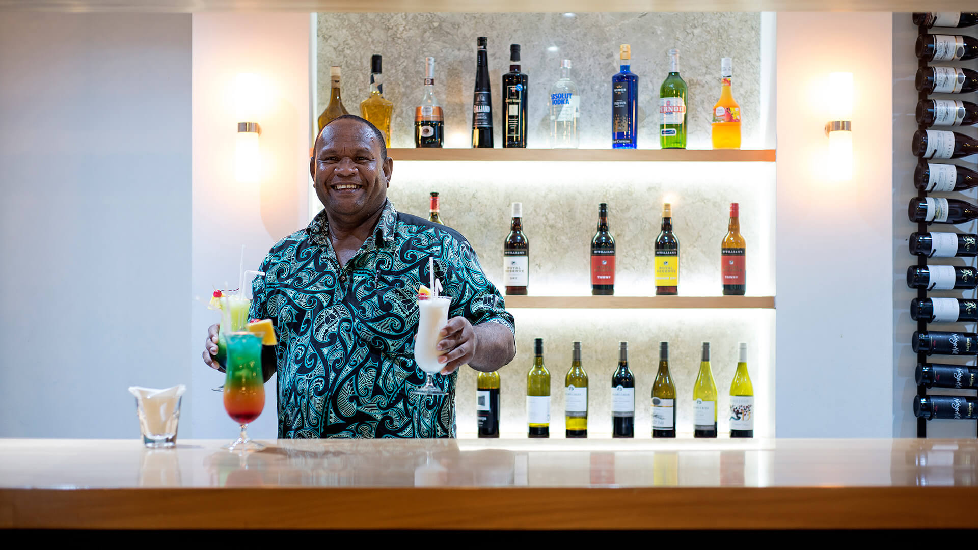 Smiling bartender at ULTIQA Fiji holding colourful tropical cocktails standing behind a well-stocked bar.