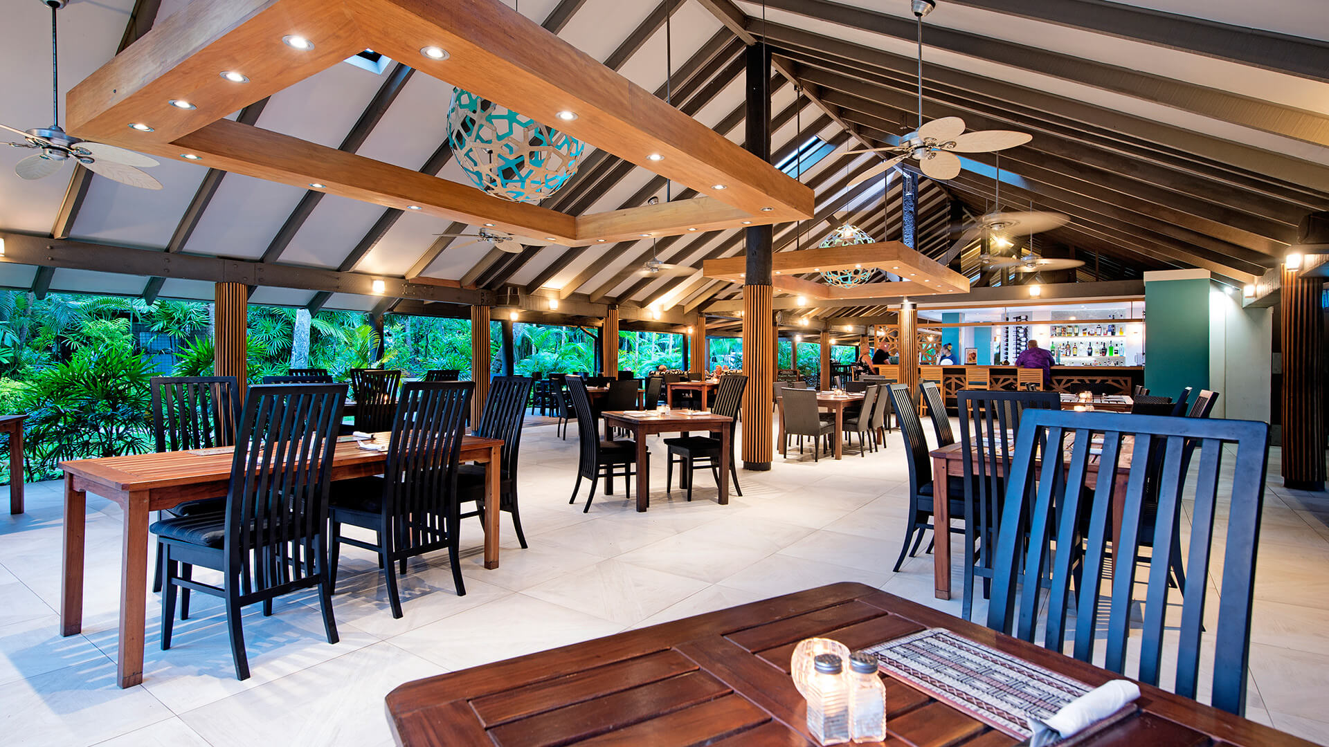 Elegant open-air dining area at ULTIQA Fiji with wooden tables, cushioned chairs, ambient lighting and lush tropical garden views.