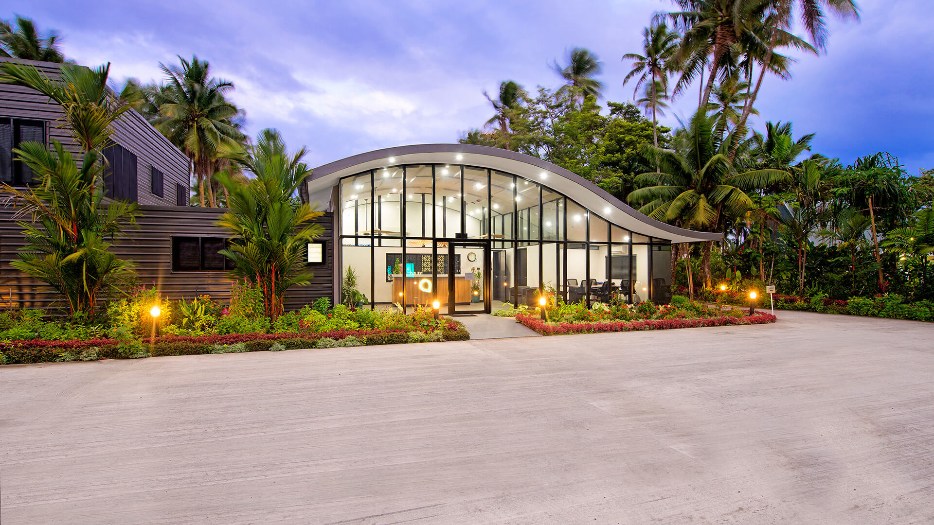 Reception building at ULTIQA Fiji with a modern curved roof, glass façade, and tropical landscaping, warmly lit at dusk.