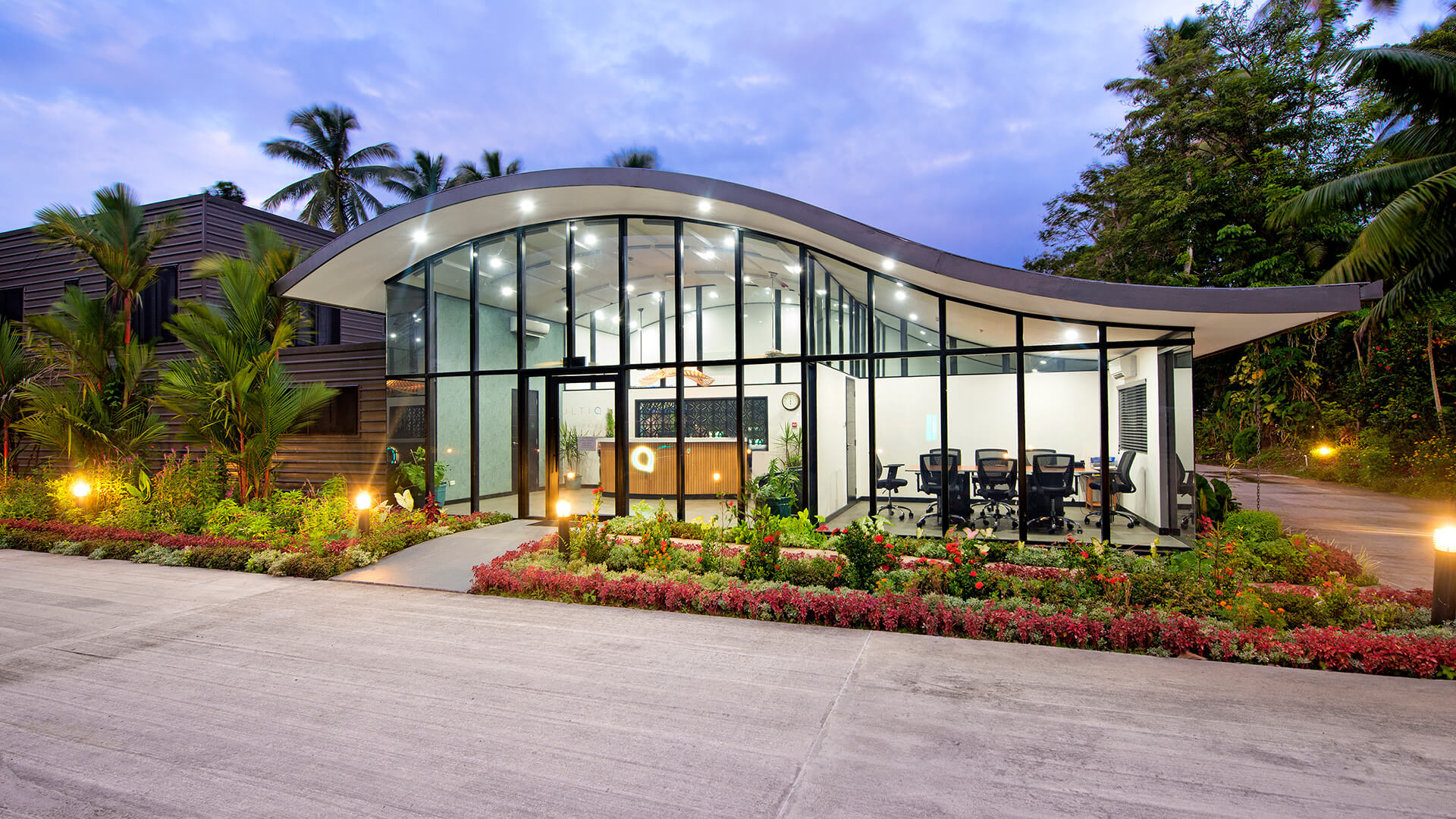 Reception area at ULTIQA Fiji with a modern curved roof design, glass walls, landscaped gardens and warm evening lighting.