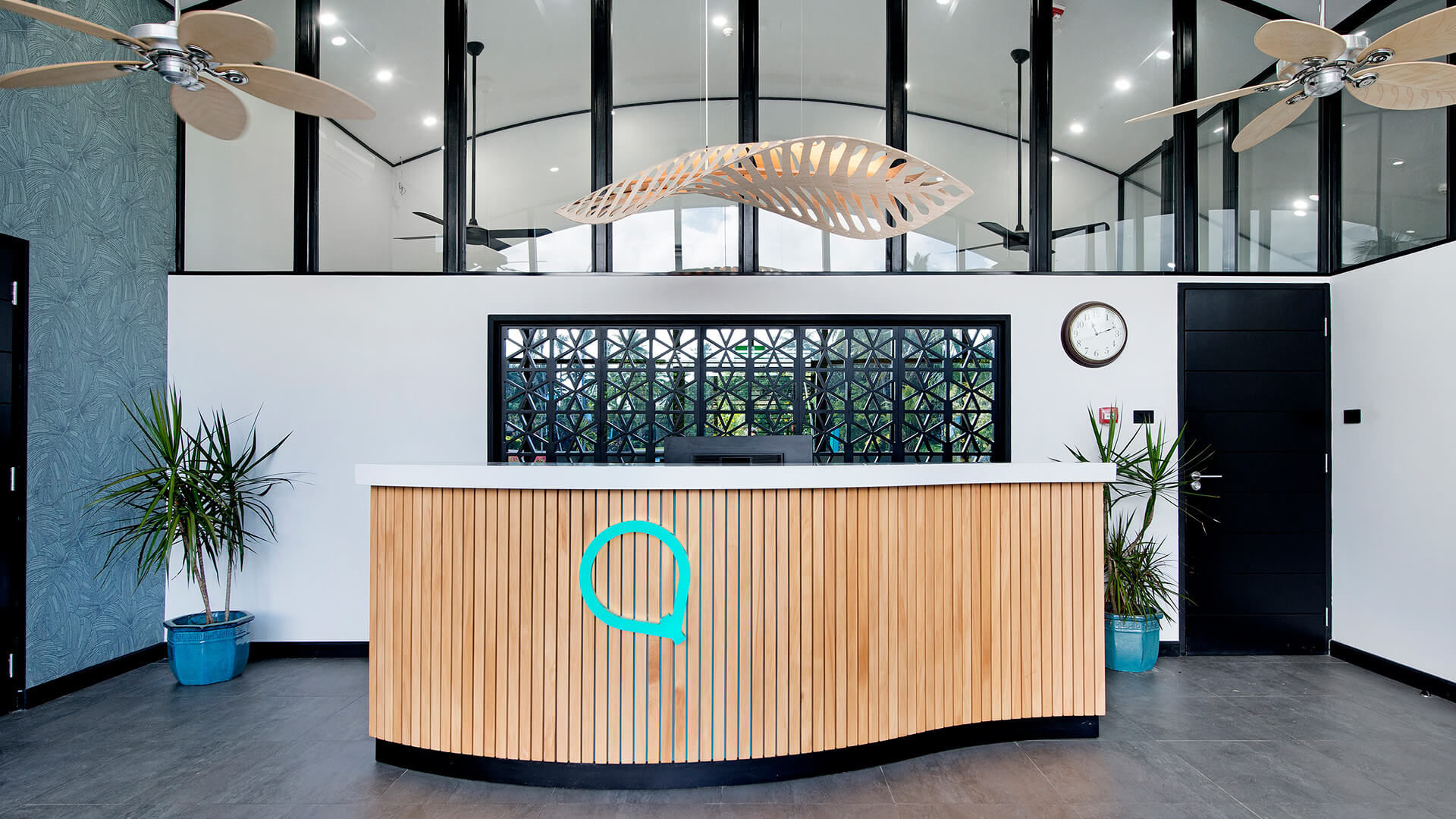 Modern reception desk at ULTIQA Fiji with a wooden slatted front, tropical plants, ceiling fans and large glass windows providing natural light.