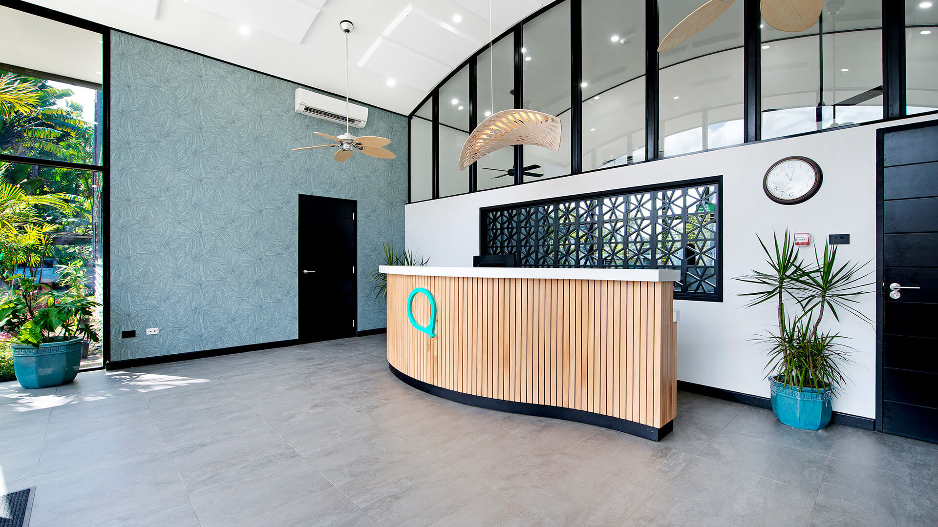 Spacious and modern reception area at ULTIQA Fiji with a curved wooden desk, tropical plants, high ceilings, large glass windows and ceiling fans.