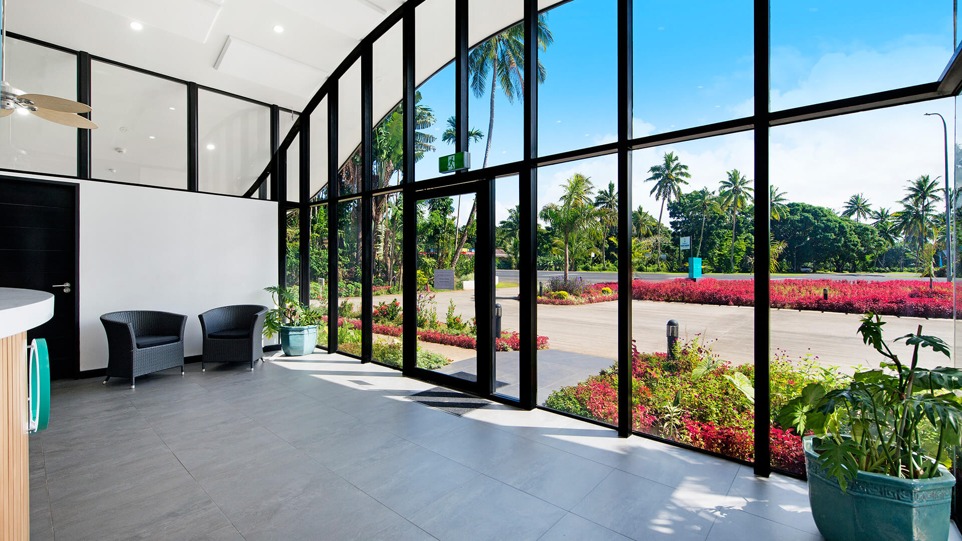Bright reception area at ULTIQA Fiji with floor-to-ceiling glass windows, tropical views, two black wicker chairs and large potted plants.