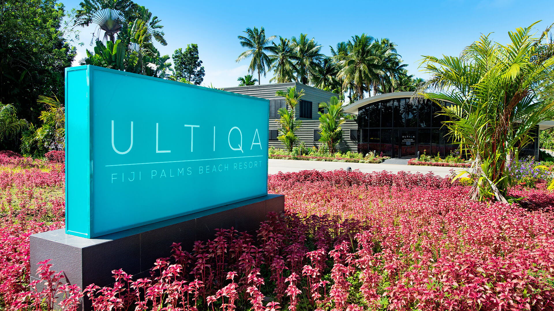 Entrance sign for ULTIQA Fiji Palms Beach Resort surrounded by vibrant pink tropical plants, with the modern reception building and palm trees in the background.