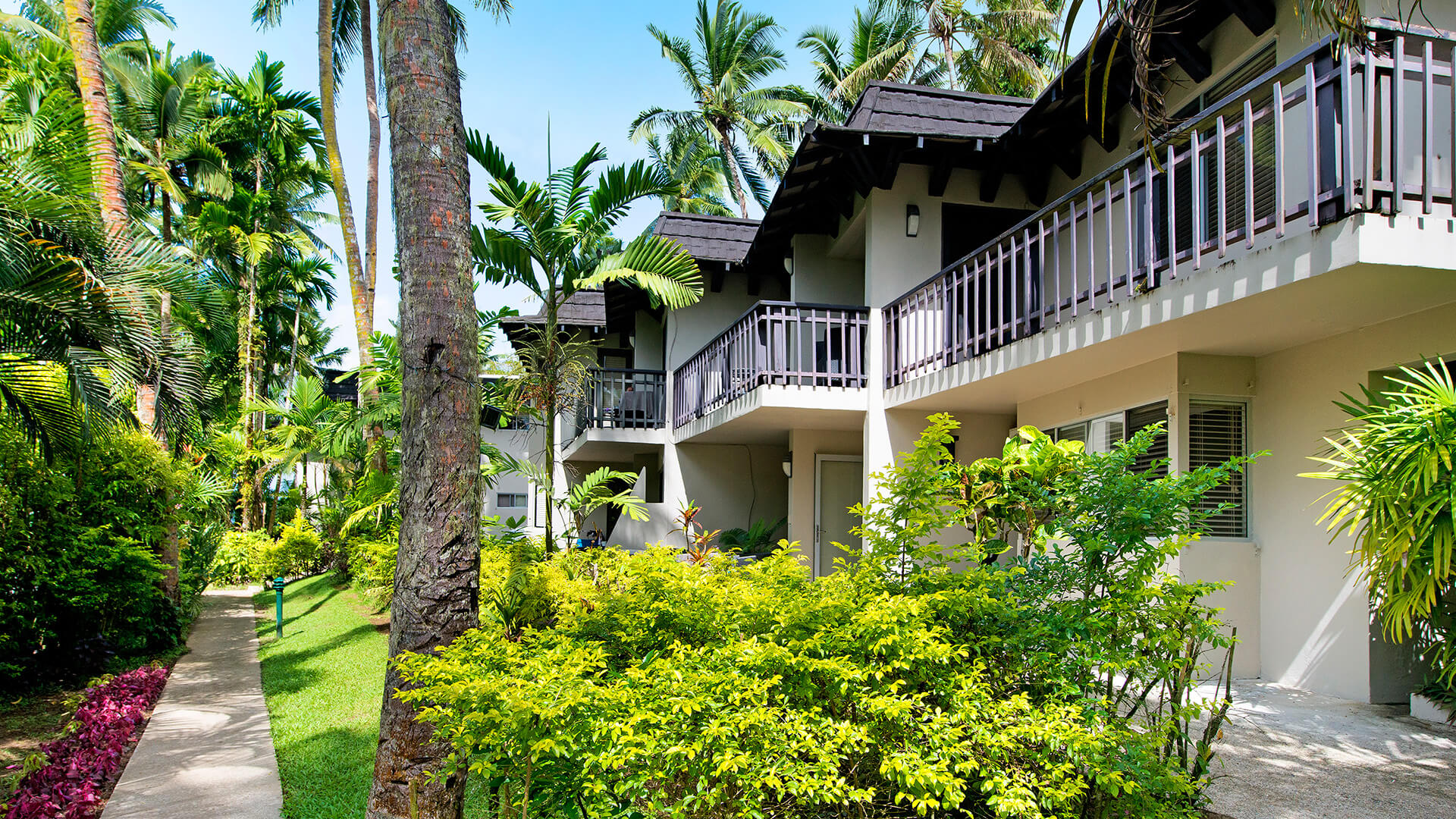 Tropical garden pathway leading to two-story accommodation units at ULTIQA Fiji Palms Beach Resort, surrounded by lush greenery and palm trees.