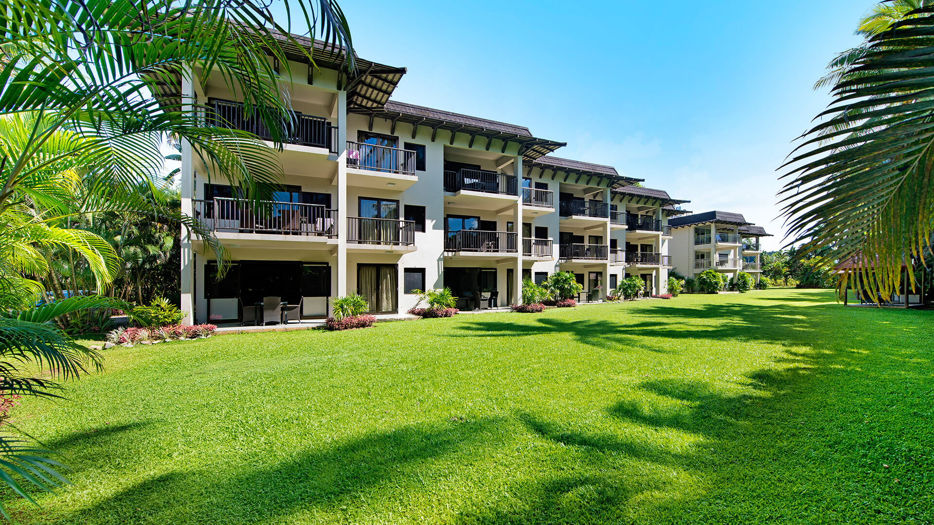 Spacious lawn area in front of multi-story accommodation buildings with private balconies at ULTIQA Fiji Palms Beach Resort, surrounded by tropical plants and palm trees.