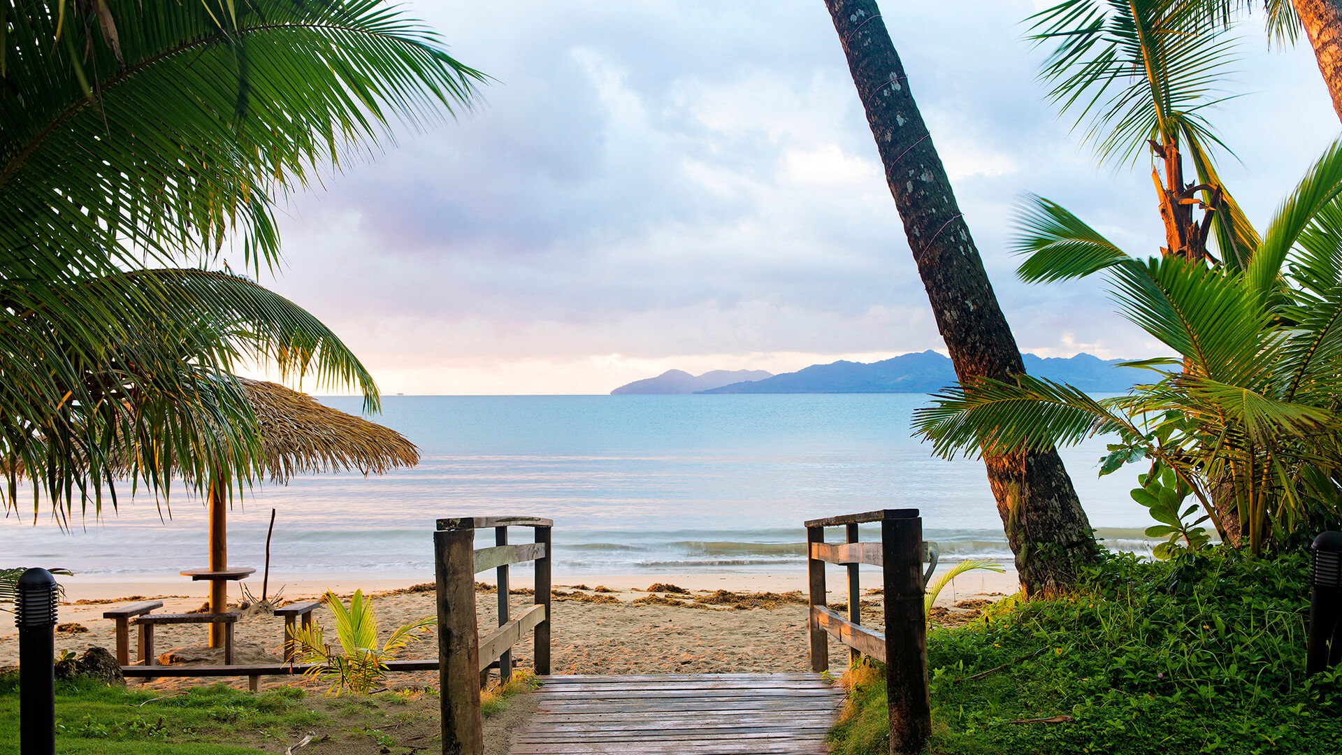Wooden walkway leading to a tranquil sandy beach with calm waters, palm trees and a thatched umbrella seating area at ULTIQA Fiji Palms Beach Resort.