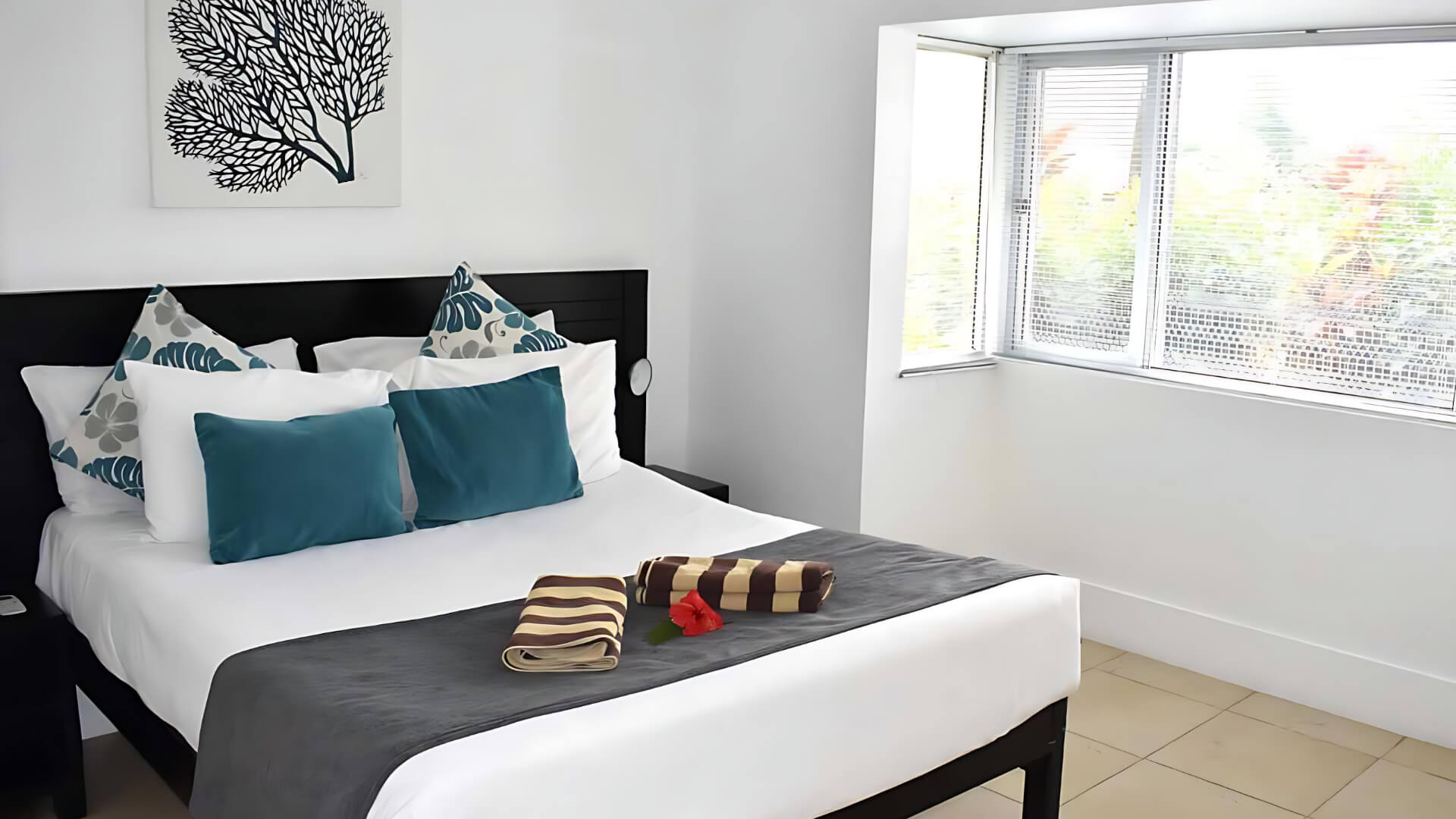 Bright bedroom in Standard 2 Bedroom 1 Bathroom Apartment at ULTIQA Fiji, featuring a queen bed with teal and patterned cushions, air conditioning, and garden views.