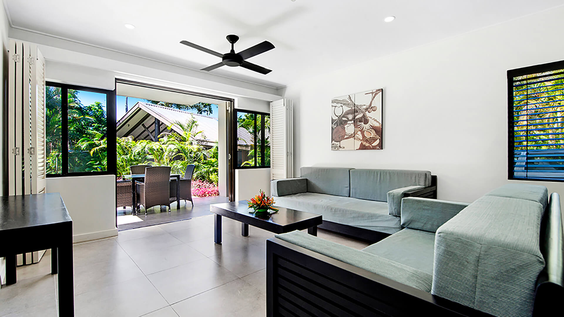 Modern living room featuring two sofas, a coffee table, and large sliding doors opening to an outdoor dining area with tropical garden views.