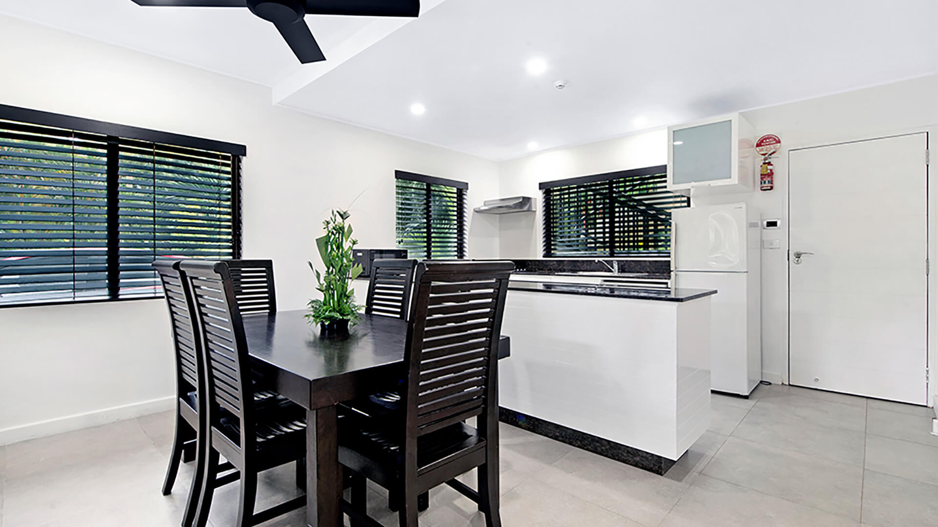 Dining area with dark wood table and six chairs next to modern kitchen with black stone benchtops in 2 Bedroom Apartment at ULTIQA Fiji Palms.