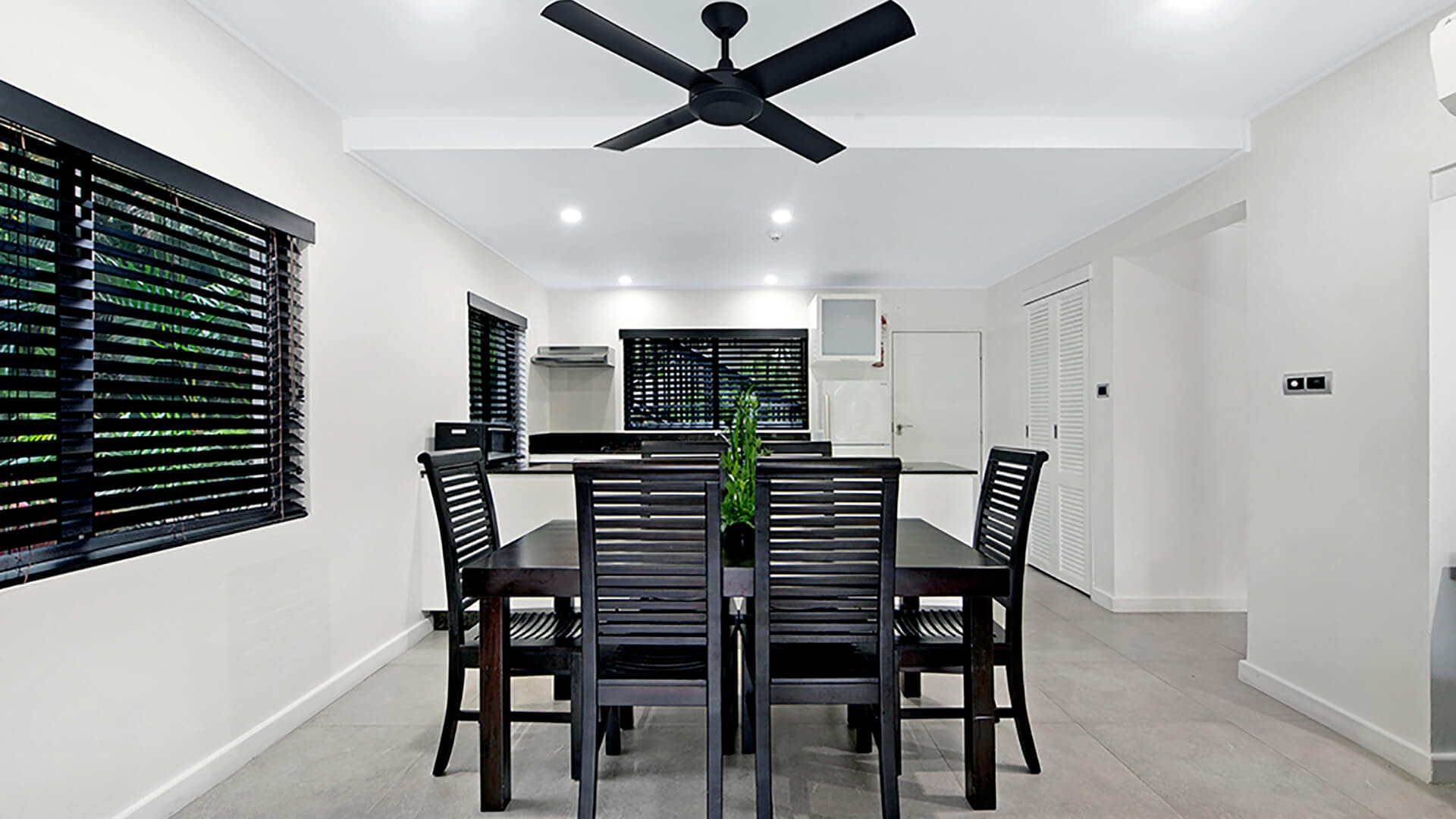 Dining area with sleek black timber table and chairs, ceiling fan, and large windows offering natural light, adjacent to the modern.