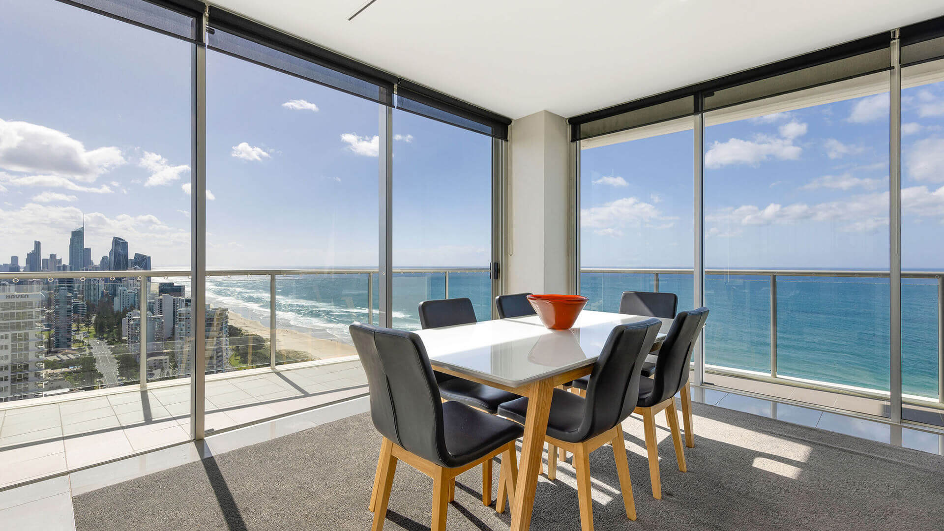 Dining area in ULTIQA Air’s 3 Bedroom Ocean View Suite with white table, black chairs, and sweeping ocean and city views through floor-to-ceiling windows.