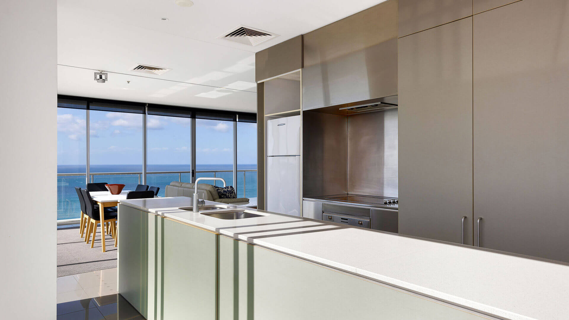Modern kitchen and dining area in ULTIQA Air’s 3 Bedroom Ocean View Suite featuring sleek cabinetry, a large island bench, and panoramic ocean views through floor-to-ceiling windows.