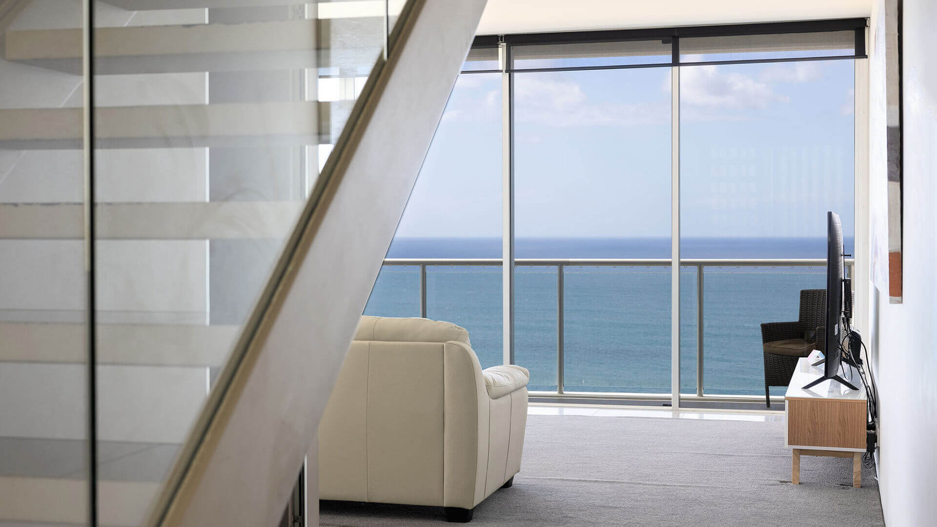 Bright living space in ULTIQA Air’s 3 Bedroom Ocean View Suite featuring a white sofa, wicker armchair, and glass-panel staircase, with sweeping ocean views through floor-to-ceiling windows.