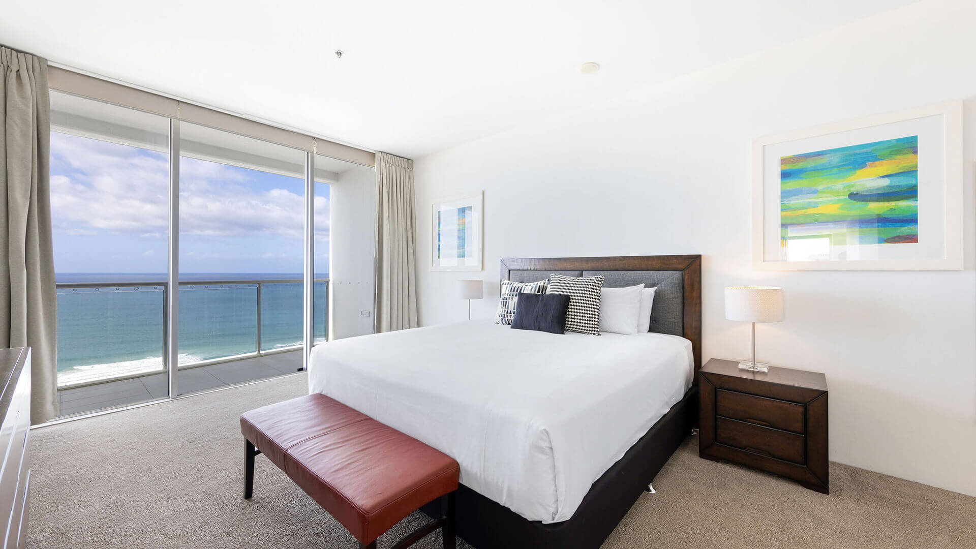 Ocean-facing bedroom in ULTIQA Air’s 3 Bedroom Ocean View Suite featuring white linens, a red bench, abstract wall art, and floor-to-ceiling windows framing panoramic coastal views.