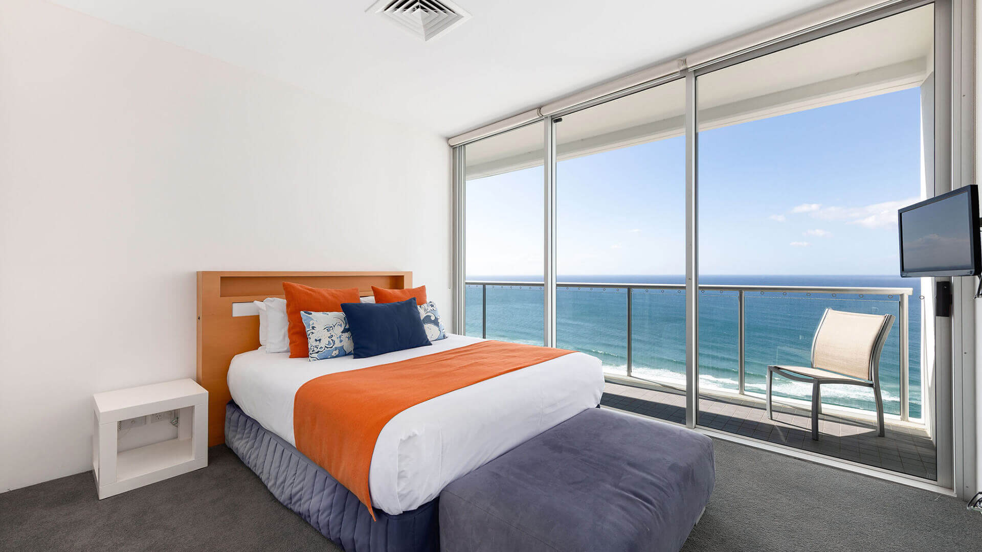 Ocean-view bedroom in ULTIQA Air’s 3 Bedroom Ocean View Suite featuring white linens, an orange throw, patterned cushions, and a private balcony with seating.