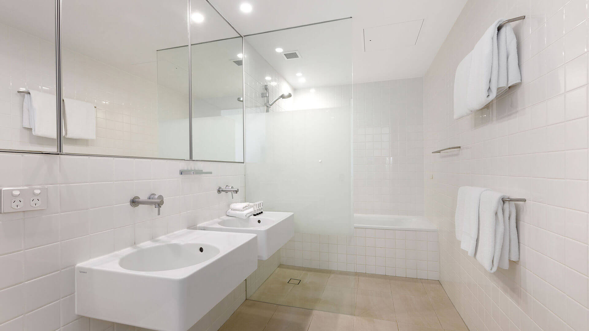 Contemporary bathroom in ULTIQA Air’s 3 Bedroom Ocean View Suite featuring dual sinks, a glass-enclosed shower, and a bathtub, with white and beige tiling throughout.