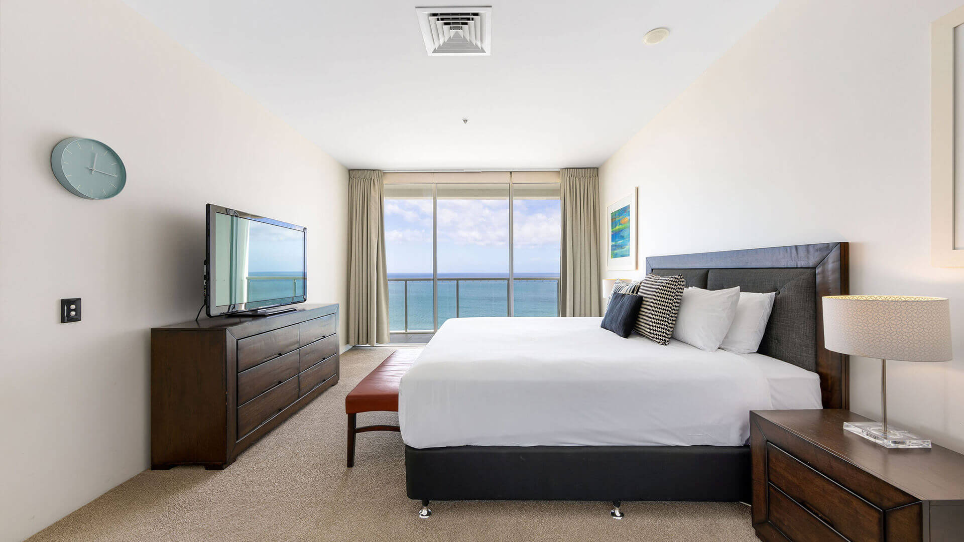 Ocean-view bedroom in ULTIQA Air’s 3 Bedroom Ocean View Suite featuring a king-sized bed with white linens, a wooden dresser with flat-screen TV, and a bench at the foot of the bed, illuminated by nat